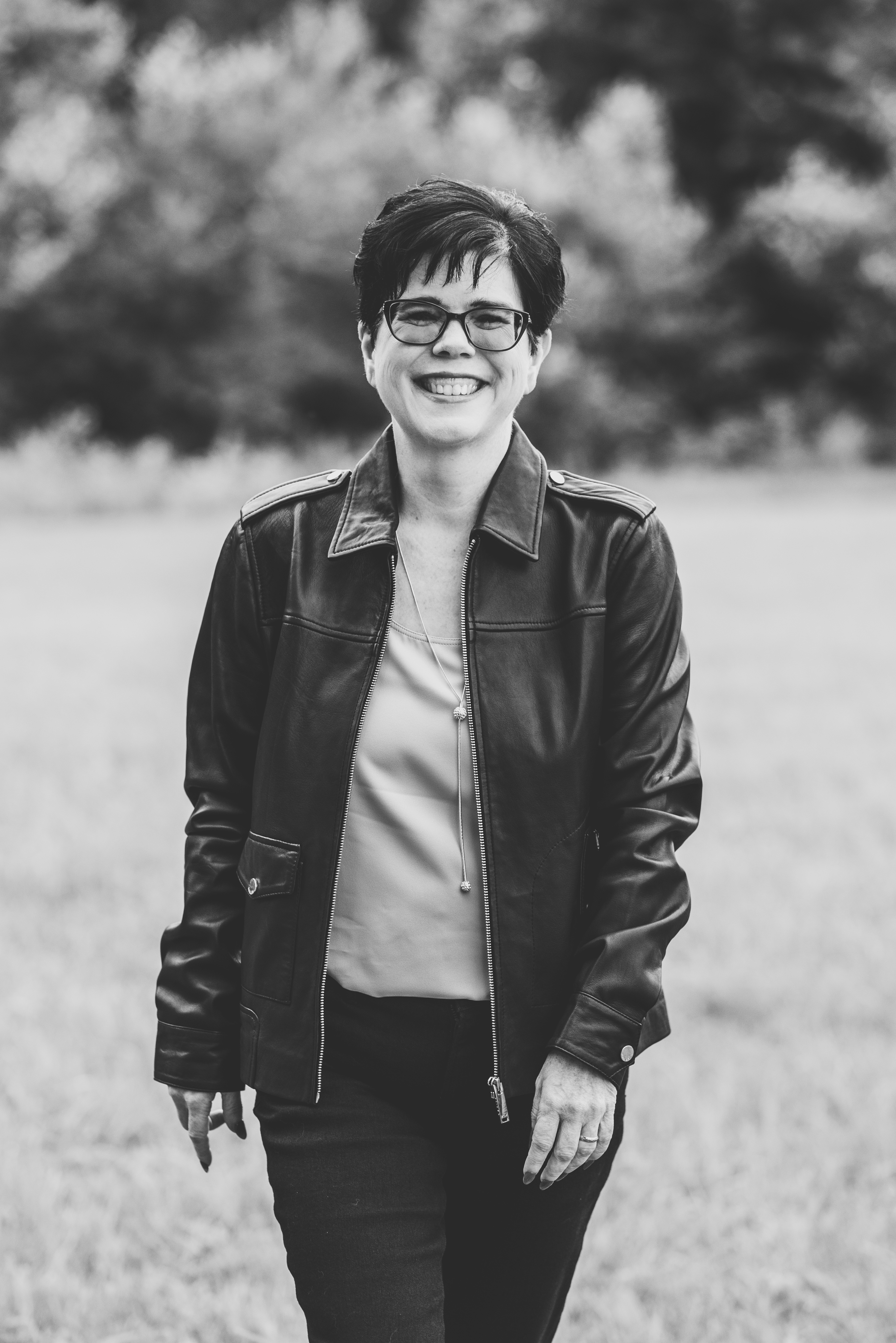 Greyscale picture of a caucasian woman with dark hair, wearing glasses, a leather jacket, a light top, and dark jeans, walking toward the camera.